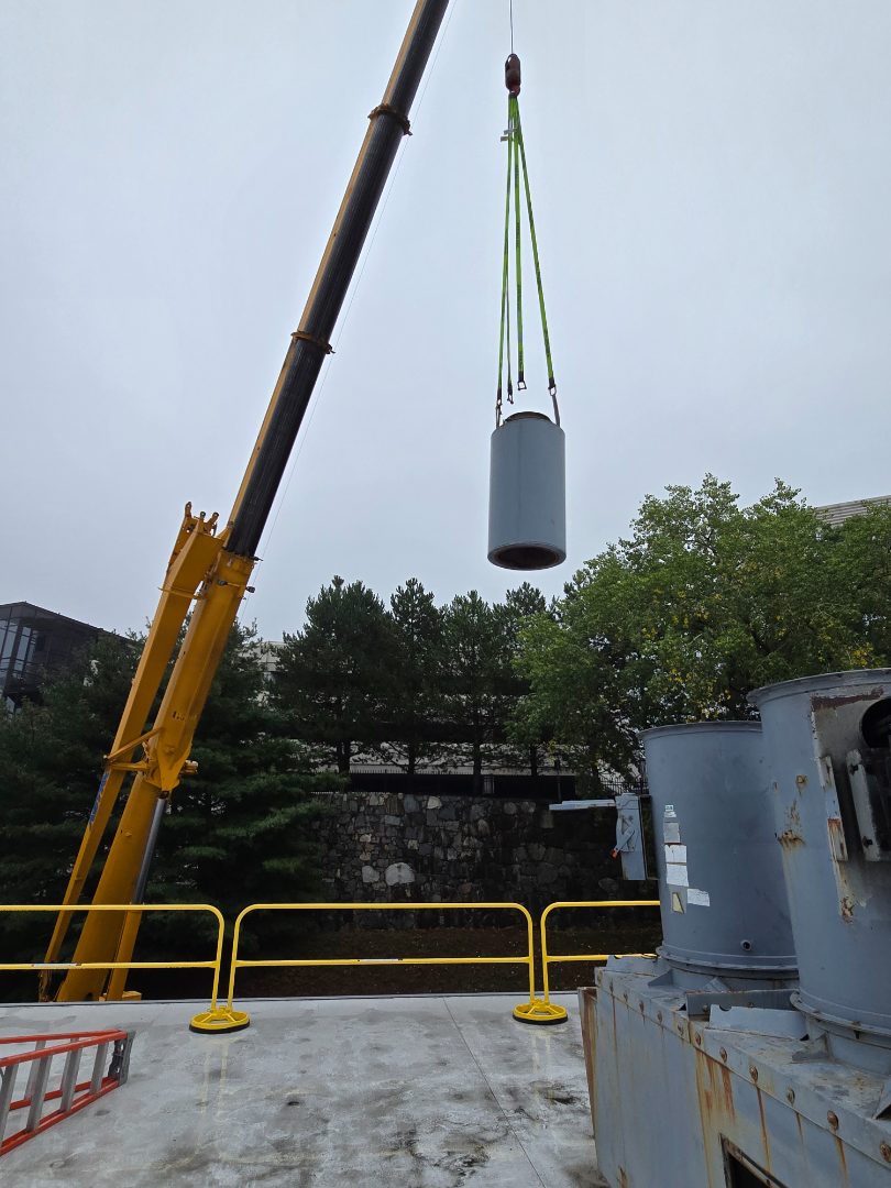 A crane lifting a tall exhaust fan stack past a stone retaining wall